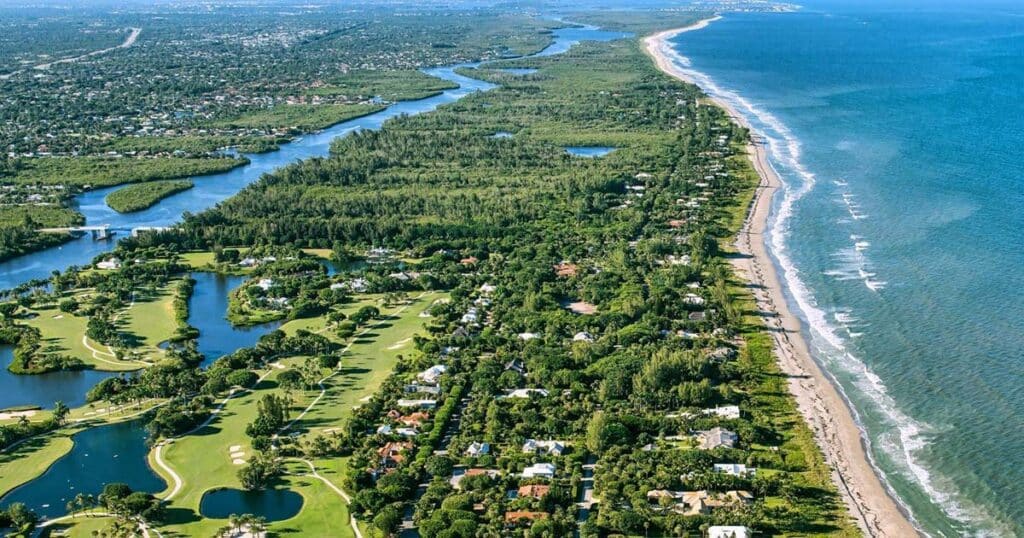 Aerial view of a scenic coastal town featuring a lush golf course, winding waterway, residential areas, and expansive beach along the ocean shoreline.