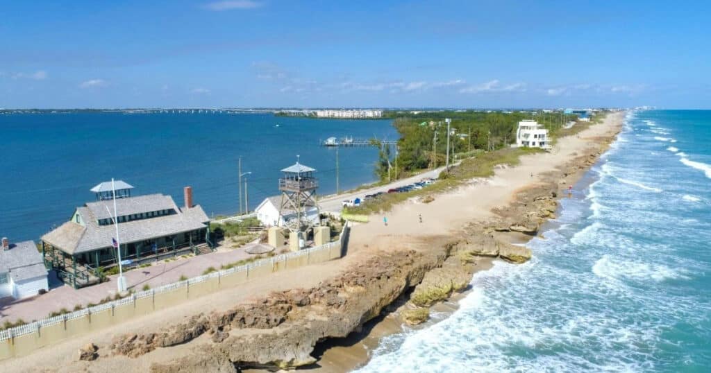 Coastal scene with rocky shoreline, waves, and beachfront houses along the ocean coast with blue sky and horizon.
